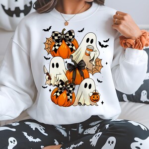 May include: White sweatshirt featuring a Halloween design with cartoon ghosts, orange pumpkins, and black bats. The pumpkins have black and white checkered bows. One ghost holds a knife, another a coffee cup, and another a jack-o'-lantern.