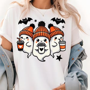 May include: White t-shirt featuring a Halloween-themed graphic. The design includes three cartoon ghosts wearing orange knit hats, holding coffee cups, and surrounded by bats and stars. The ghosts have smiling faces and are holding coffee cups.