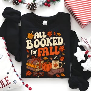 Puede incluir: Sudadera negra con el texto "ALL BOOKED for FALL" en crema y naranja. El dise&ntilde;o incluye hojas de oto&ntilde;o, una calabaza, libros, una taza de caf&eacute; y gafas. La sudadera est&aacute; rodeada de decoraciones.