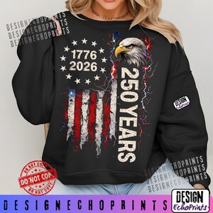 Puede incluir: Sudadera negra con un dise&ntilde;o de bandera estadounidense desgastada, el texto "1776 2026" y "250 YEARS". El dise&ntilde;o incluye una cabeza de &aacute;guila y rayos. La sudadera tiene mangas largas.