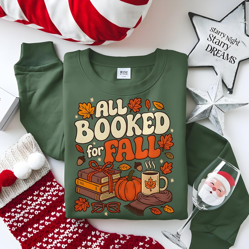Puede incluir: Sudadera verde con el texto "ALL BOOKED for FALL" en naranja y crema, con gr&aacute;ficos tem&aacute;ticos de oto&ntilde;o. La imagen incluye una calabaza, libros, una taza de caf&eacute; y hojas de oto&ntilde;o. La sudadera se combina con pantalones deportivos verdes y decoraciones navide&ntilde;as.