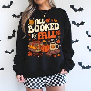 Puede incluir: Sudadera negra con el texto "All Booked for Fall" en naranja y crema. El dise&ntilde;o incluye libros, una calabaza, una taza con una hoja y hojas de oto&ntilde;o. La sudadera se combina con pantalones cortos a cuadros blancos y negros.