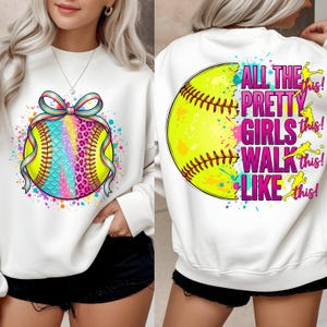 Puede incluir: Sudadera blanca con diseño de softbol. La parte delantera tiene un softbol con un patrón de escamas arcoíris y un lazo. La parte trasera muestra un softbol con el texto "ALL THE PRETTY GIRLS WALK LIKE THIS!"