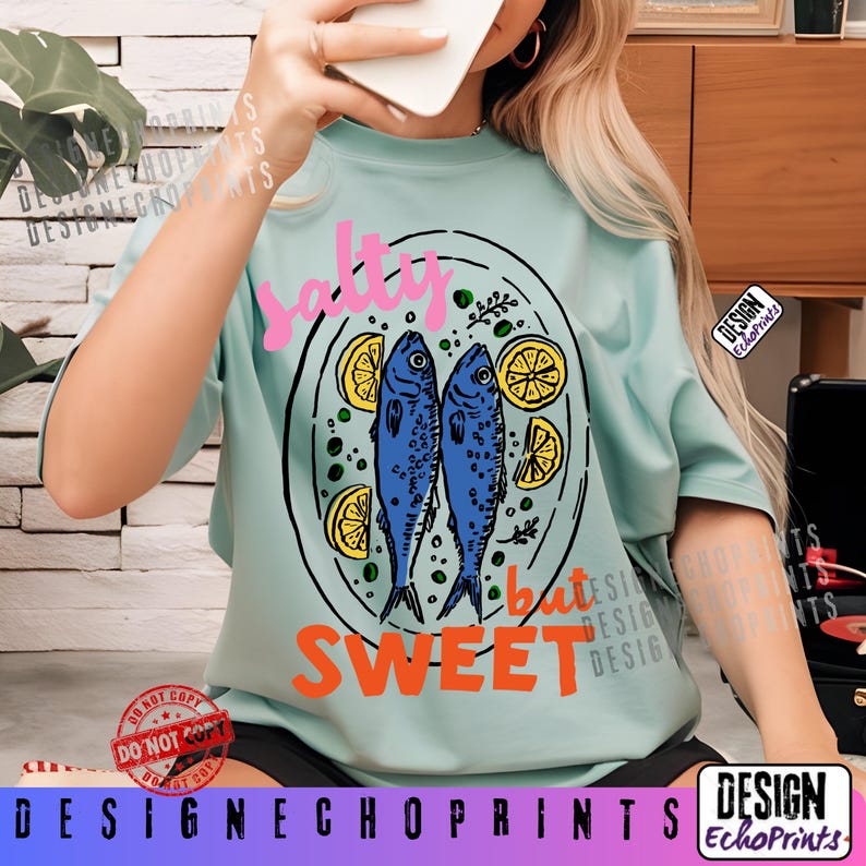 Puede incluir: Camiseta azul claro con un gr&aacute;fico de dos peces azules, rodajas de lim&oacute;n y las palabras "salty but sweet" en rosa y naranja. La camiseta tiene mangas cortas y cuello redondo. El dise&ntilde;o est&aacute; centrado en la parte delantera.