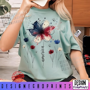 K&ouml;nnte beinhalten: Hellblaues T-Shirt mit einem Blumendesign in Rot, Wei&szlig; und Blau, mit dem Wort "america" in Schreibschrift. Das Design umfasst Sterne, Schmetterlinge und kleinere Blumen. Das T-Shirt hat kurze &Auml;rmel.