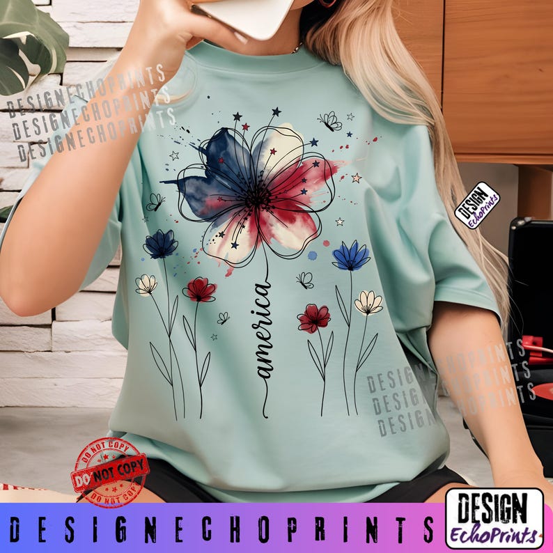 K&ouml;nnte beinhalten: Hellblaues T-Shirt mit einem Blumendesign in Rot, Wei&szlig; und Blau, mit dem Wort "america" in Schreibschrift. Das Design umfasst Sterne, Schmetterlinge und kleinere Blumen. Das T-Shirt hat kurze &Auml;rmel.
