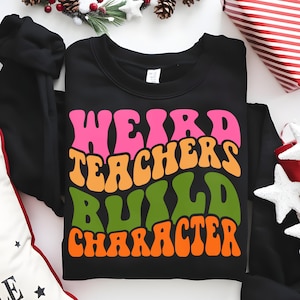 Puede incluir: Sudadera negra con el texto "WEIRD TEACHERS BUILD CHARACTER" en letras rosas, naranjas, verdes y naranjas. La sudadera está doblada sobre una superficie blanca con adornos navideños.