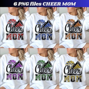 May include: White sweatshirt featuring the text "Cheer Mom" in a bold, black font with a pom-pom and megaphone graphic. The pom-poms and text have a leopard print design in various colors, including red, blue, orange, purple, green, and yellow.
