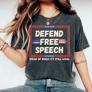 Puede incluir: Camiseta gris oscuro con las palabras "DEFEND FREE SPEECH" en blanco, rojo y azul. El diseño incluye una bandera estadounidense y la frase "SPEAK UP WHILE IT'S STILL LEGAL".