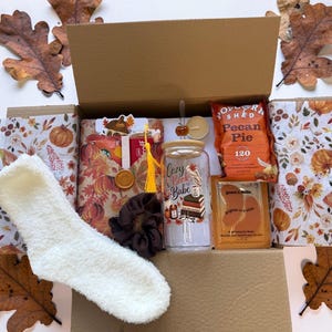 May include: A gift box containing a white fuzzy sock, a brown scrunchie, a glass tumbler with a bamboo lid and straw featuring the text Cozy Book Babe, a bag of Pecan Pie popcorn, and a gold-toned eye mask packet, all nestled in pumpkin-print paper.