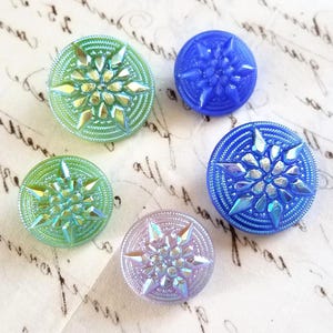 May include: Five round glass buttons featuring a raised starburst or snowflake pattern. The collection includes two green iridescent buttons, two blue buttons with varying finishes, and one light purple iridescent button with textured edges.