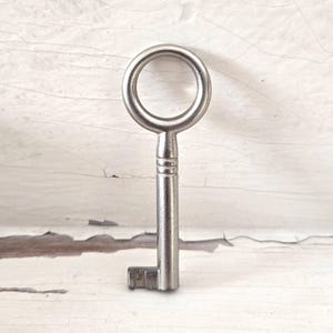 May include: A silver-toned metal skeleton key featuring a circular bow, a cylindrical shank with two horizontal decorative grooves near the top, and a simple, rectangular bit at the bottom end of the shaft, standing in an upright position.