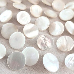 May include: A collection of round, white mother-of-pearl buttons scattered across the frame. Each button features a smooth, iridescent surface with natural variations in luster and tone, showcasing the characteristic sheen of shell material.