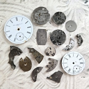 May include: A collection of vintage watch parts including two white circular watch faces with black Roman numerals and sub-dials, various metallic gears, plates, and bridges in silver and brass tones, showing signs of age and mechanical wear.