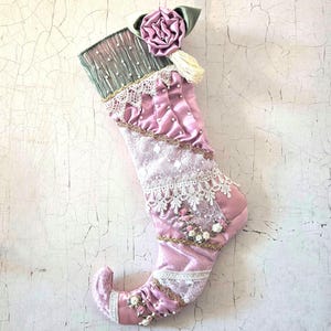 May include: A decorative Christmas stocking in shades of dusty pink, featuring a ruffled cuff with a sage green ribbon rose, white lace trim, gold braided accents, and small floral embellishments. The stocking has a curved toe and a hanging loop.