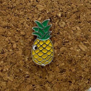 May include: A yellow enamel pineapple pin with green leaves and a white eye. The pin is on a cork board.