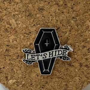 Peut inclure: Épinglette en forme de cercueil en émail noir avec une bannière blanche qui dit "Let's Hide". Le cercueil a une petite croix sur le dessus.