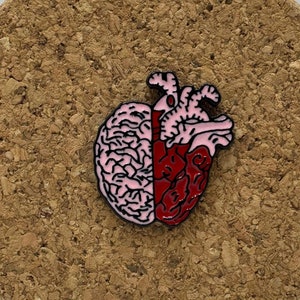 Puede incluir: Un pin de esmalte rosa y rojo que representa un cerebro y un corazón fusionados. El cerebro está en el lado izquierdo y el corazón en el lado derecho.