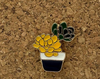 May include: A gold-coloured enamel pin featuring a potted succulent plant. The succulent is green and yellow, and the pot is white with a black rim.