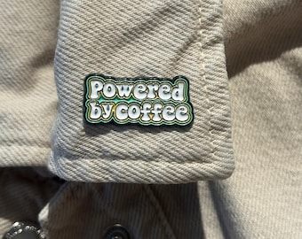 Könnte beinhalten: Nahaufnahme einer beigefarbenen Jeansjacke mit einer kleinen Emaille-Anstecknadel am Kragen. Auf der Nadel steht "Powered by coffee" in weißer und grüner Schrift. Die Jacke hat silberne Knöpfe und eine strukturierte Oberfläche.