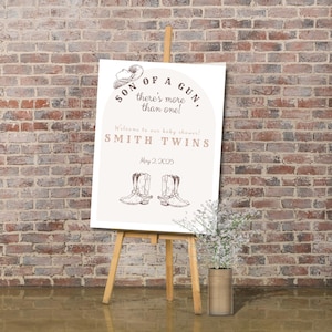 May include: A white sign with brown text and illustrations announcing a baby shower for twins. The sign reads "Son of a Gun, there's more than one!" and "Smith Twins" with the date "May 2, 2025". The sign is on an easel with a vase of white flowers in front of a brick wall.