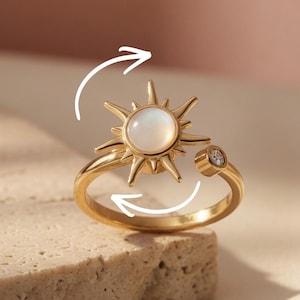Könnte beinhalten: Ein goldfarbener Ring mit einem Sonnenmuster, einem zentralen weißen Stein und einem kleinen Diamanten. Der Ring wird auf einer strukturierten, hellfarbigen Oberfläche vor einem sanften rosa Hintergrund präsentiert. Das Sonnenmuster ist das Hauptmerkmal.