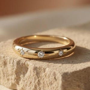Könnte beinhalten: Ein goldener Ring mit drei kleinen, klaren Edelsteinen. Der Ring liegt auf einer rauen, beigefarbenen Steinoberfläche, mit einem weichen, neutralen Hintergrund.