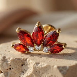May include: A gold-toned jewelry piece featuring a floral design with red gemstone petals and small white accent stones. The jewelry is set on a textured, light-colored surface, with a soft background.