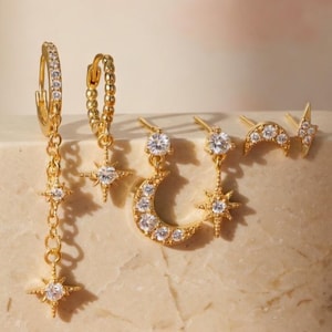 Pode incluir: Uma coleção de brincos dourados com designs celestiais. Os brincos incluem estrelas, luas crescentes e raios, todos adornados com pedras preciosas claras e brilhantes. Os brincos são exibidos em um bloco texturizado branco sujo.