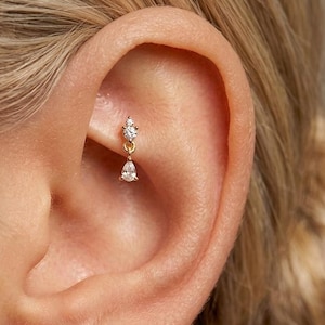 Puede incluir: Un pendiente dorado con una pequeña gema brillante en la parte superior y una gema en forma de lágrima colgando. El pendiente está engarzado en una oreja, mostrando el diseño y el brillo. Una joya delicada.