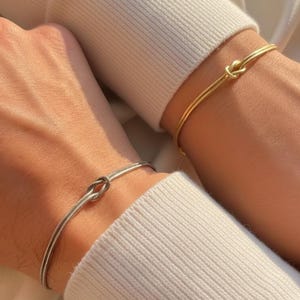 Puede incluir: Dos pulseras, una plateada y otra dorada, cada una con un diseño de nudo. Las pulseras son finas y delicadas, con una estética simple pero elegante. La imagen muestra las pulseras en muñecas, lo que sugiere un enfoque en joyas y accesorios.