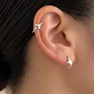 Puede incluir: Primer plano de una oreja adornada con dos pendientes plateados en forma de estrella. Uno es un brazalete y el otro un perno. La joyería es minimalista y moderna, reflejando un tema celestial.