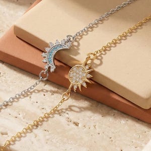 May include: Two delicate bracelets, one silver and one gold, each with celestial charms. The silver bracelet has a crescent moon with blue accents, and the gold one has a sunburst design. Both bracelets are displayed on a neutral background.