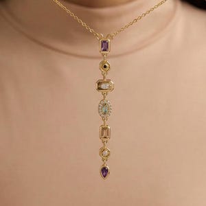 Può includere: Una collana color oro con un pendente verticale con varie pietre preziose nei colori viola, verde e trasparente. La collana è presentata da una persona.