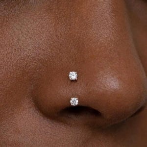 May include: A close-up of a nose featuring two small diamond nose studs. The studs are set in a minimalist style, adding a subtle sparkle. The skin tone is a warm brown, creating a contrast with the bright gems. Ideal for everyday wear.