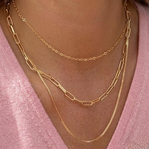 May include: A layered gold-tone necklace set consisting of three distinct chains. The top chain is a delicate cable link, the middle is a bold elongated paperclip link, and the bottom is a fine box chain, all worn together against a pink fabric.