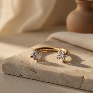 May include: A gold ring with two large, clear gemstones. The ring is set on a light-colored stone surface, with a small brown vase and draped fabric in the background. The ring's design features a curved band with the gemstones set at each end.