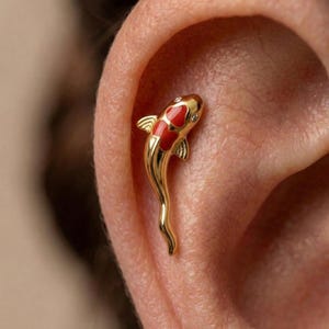 Può includere: Un orecchino a forma di pesce koi dorato con dettagli in smalto rosso e bianco e piccole pietre trasparenti. L'orecchino è a lobo e si indossa nell'orecchio. Il design del pesce è dettagliato ed elegante.