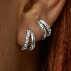 Può includere: Primo piano di due orecchini a cerchio argentati. Uno è un cerchio singolo, l'altro un doppio cerchio. Entrambi sono incastonati con piccoli cristalli scintillanti. Gli orecchini sono su uno sfondo sfocato.