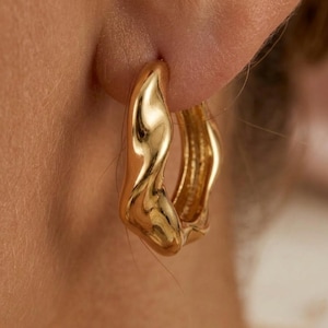May include: Close-up of a pair of gold-colored hoop earrings. The earrings have a unique, organic shape with a smooth, reflective surface. The design appears to be a modern take on a classic style, suitable for everyday wear or special occasions.