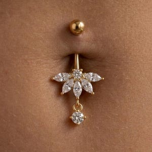 Puede incluir: Un piercing para el ombligo de color dorado con un diseño floral con piedras de circonita cúbica transparentes, de talla marquesa y redonda. La bola superior también es dorada.