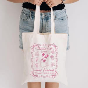 May include: Cream-colored tote bag with a pink graphic. The graphic includes the text "Happily Ever After" and "Ashley's Bachelorette DISNEY WORLD FL". The design features Minnie Mouse and other themed illustrations.
