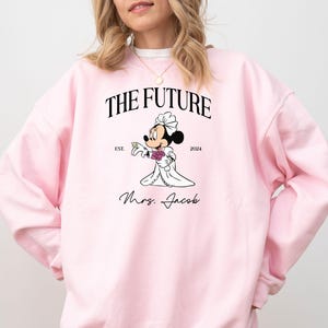 May include: Pink sweatshirt with a graphic of a cartoon bride Mickey Mouse with the text "The Future Est. 2024 Mrs. Jacob"