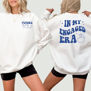 May include: Two white sweatshirts with blue text. One sweatshirt says "Future Mrs. Martinez" and the other says "In My Engaged Era" with a diamond ring graphic.