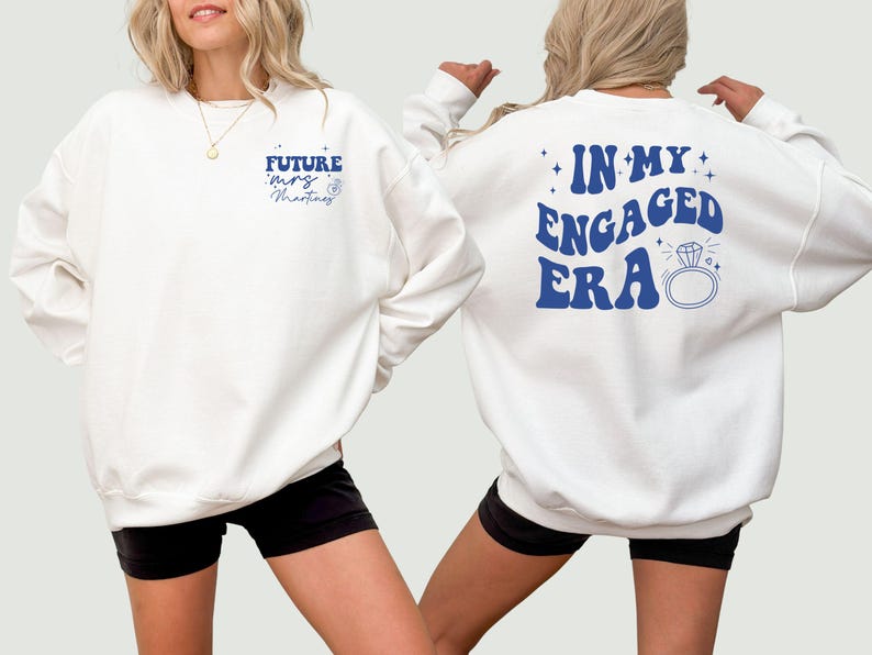May include: Two white sweatshirts with blue text. One sweatshirt says "Future Mrs. Martinez" and the other says "In My Engaged Era" with a diamond ring graphic.