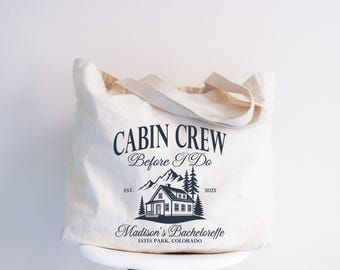 May include: A cream-coloured tote bag featuring the text "CABIN CREW Before I Do" in dark blue. A mountain and cabin graphic is below the text, with "Madison's Bachelorette ESTES PARK, COLORADO" at the bottom.