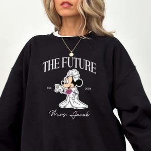 May include: Black sweatshirt with a white graphic of Mickey Mouse dressed as a bride. The text "The Future" is above Mickey Mouse and "Est. 2024" is below. The text "Mrs. Jacob" is below Mickey Mouse.