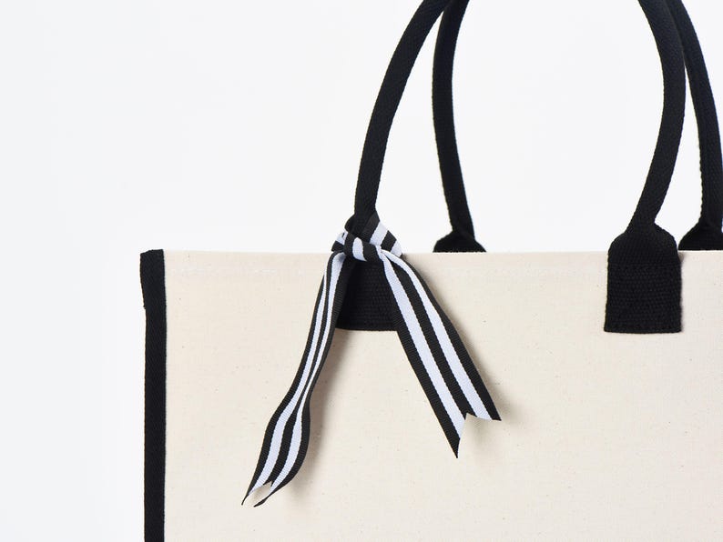 Op de afbeelding: Een witte canvas tote bag met zwarte bies en zwarte handvatten. Een zwart-wit gestreept lint is om de handvatten gebonden.