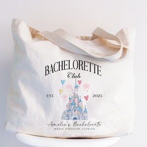 May include: A white canvas tote bag with black text that reads "Bachelorette Club Est. 2025" and a graphic of Cinderella's castle with balloons. The text below the graphic reads "Amelia's Bachelorette Magic Kingdom, Florida."