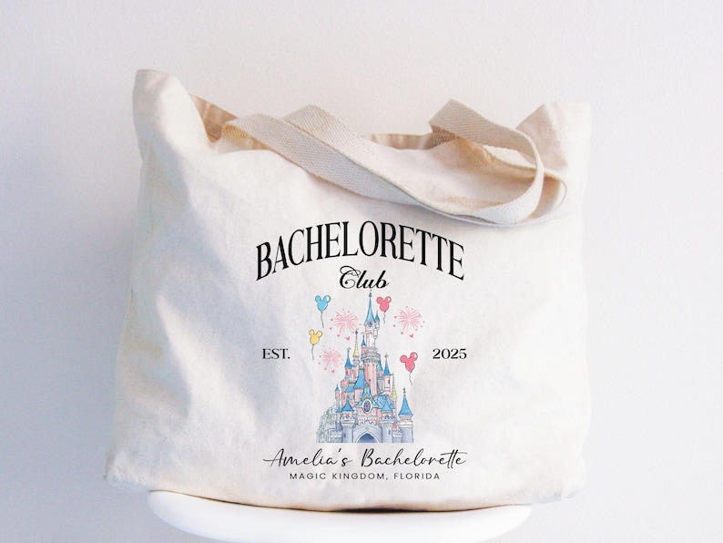 May include: A white canvas tote bag with black text that reads "Bachelorette Club Est. 2025" and a graphic of Cinderella's castle with balloons. The text below the graphic reads "Amelia's Bachelorette Magic Kingdom, Florida."